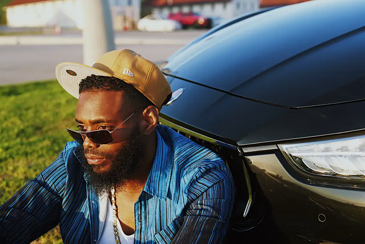 Close-up of KingJu1c3 in a tan cap and aviators, leaning against a black Maserati