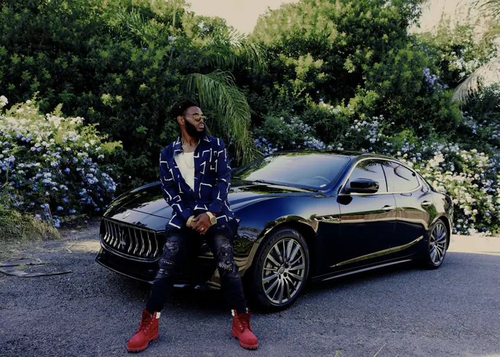KingJu1c3 sitting on the hood of a black Maserati in front of flowering hedges