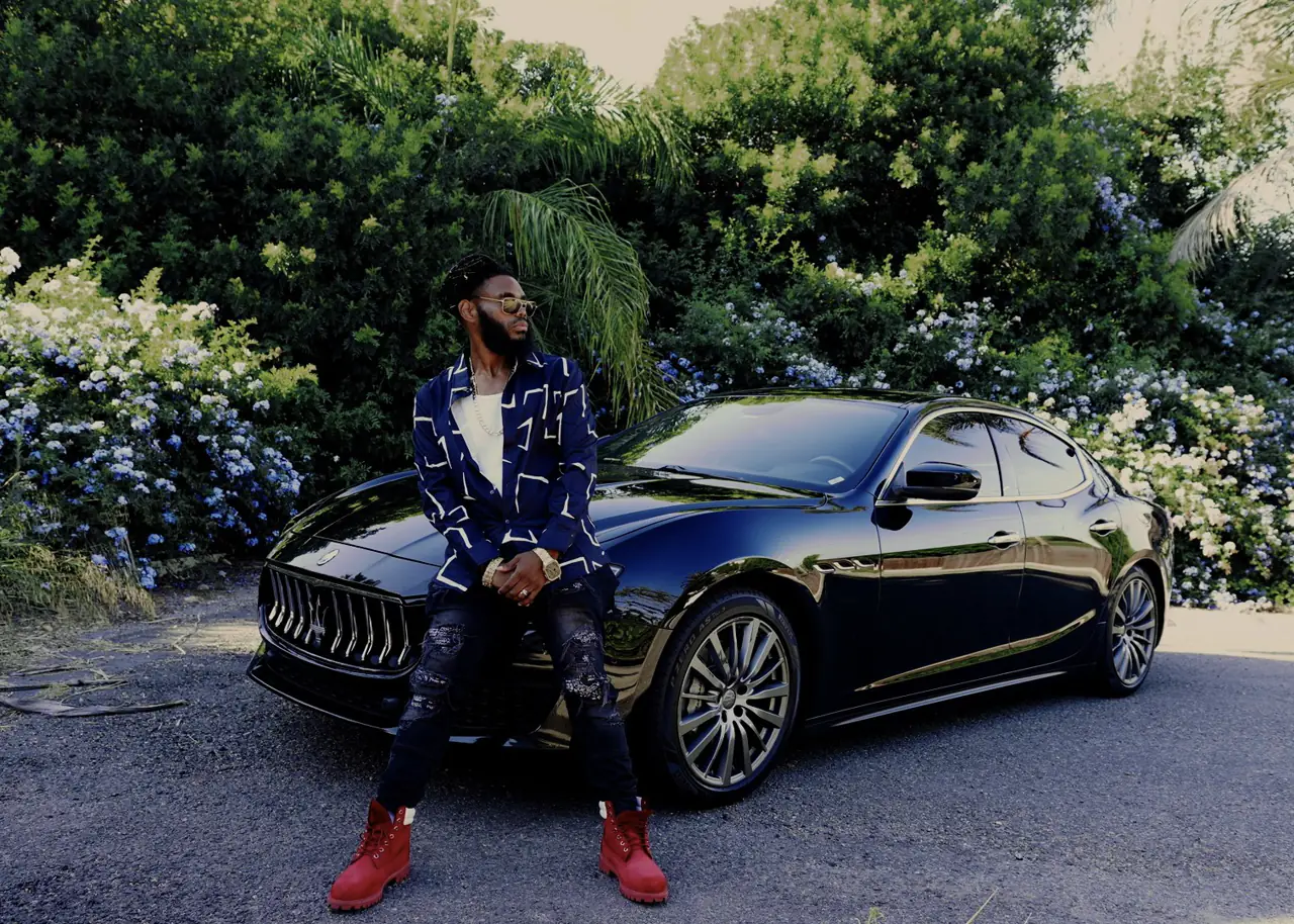 KingJu1c3 sitting on the hood of a black Maserati in front of flowering hedges, red boots