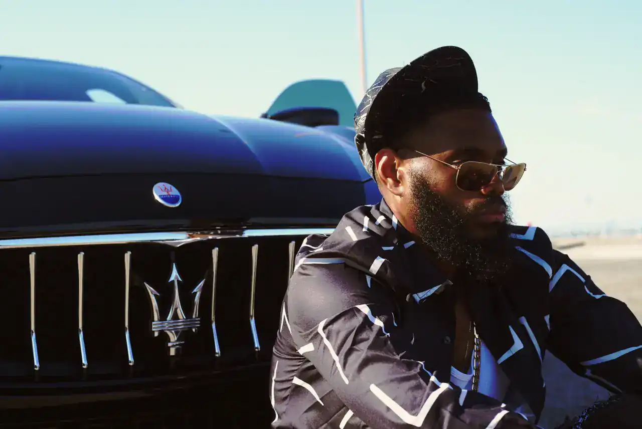 KingJu1c3 in profile beside the Maserati grille, wearing a black and white shirt and cap