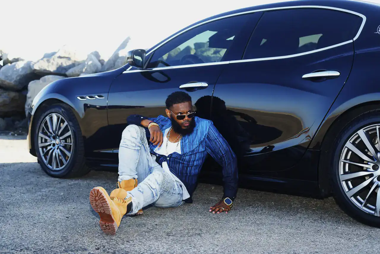 KingJu1c3 seated on the ground against a black Maserati, glancing over the shoulder