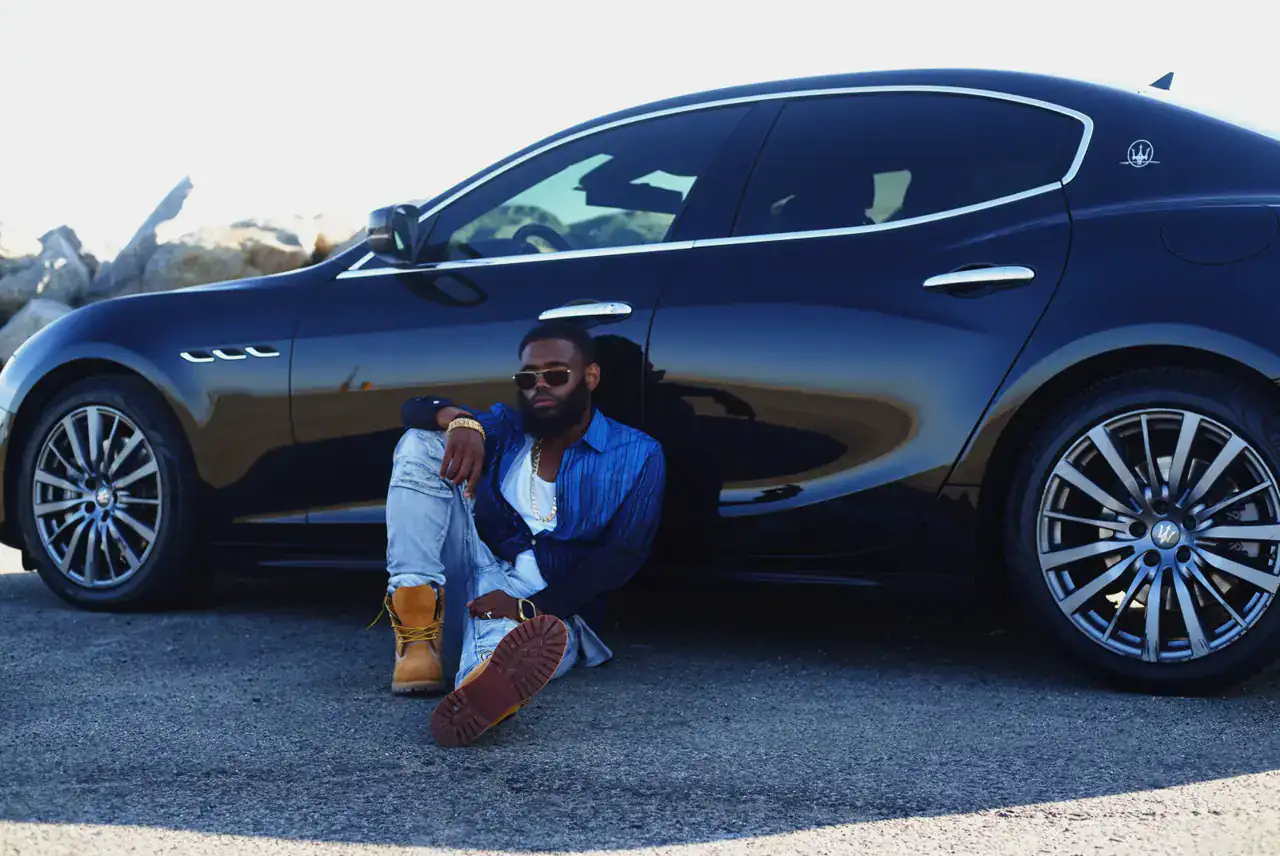 KingJu1c3 seated against the side of a black Maserati beside a rock wall