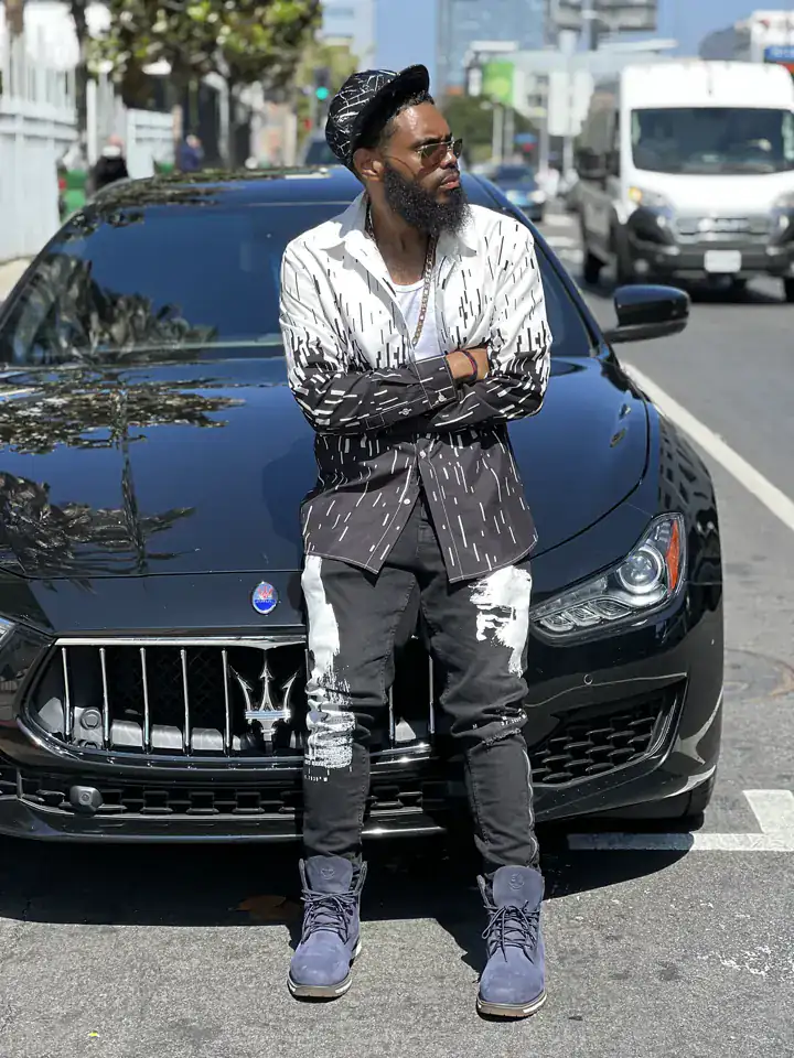 KingJu1c3 leaning on a black Maserati on a city street, arms crossed