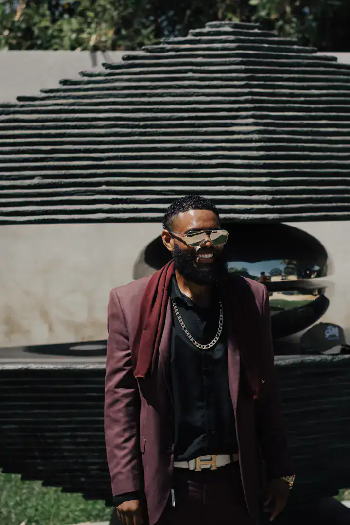 KingJu1c3 smiling in a burgundy blazer and gold chain in front of a stone sculpture