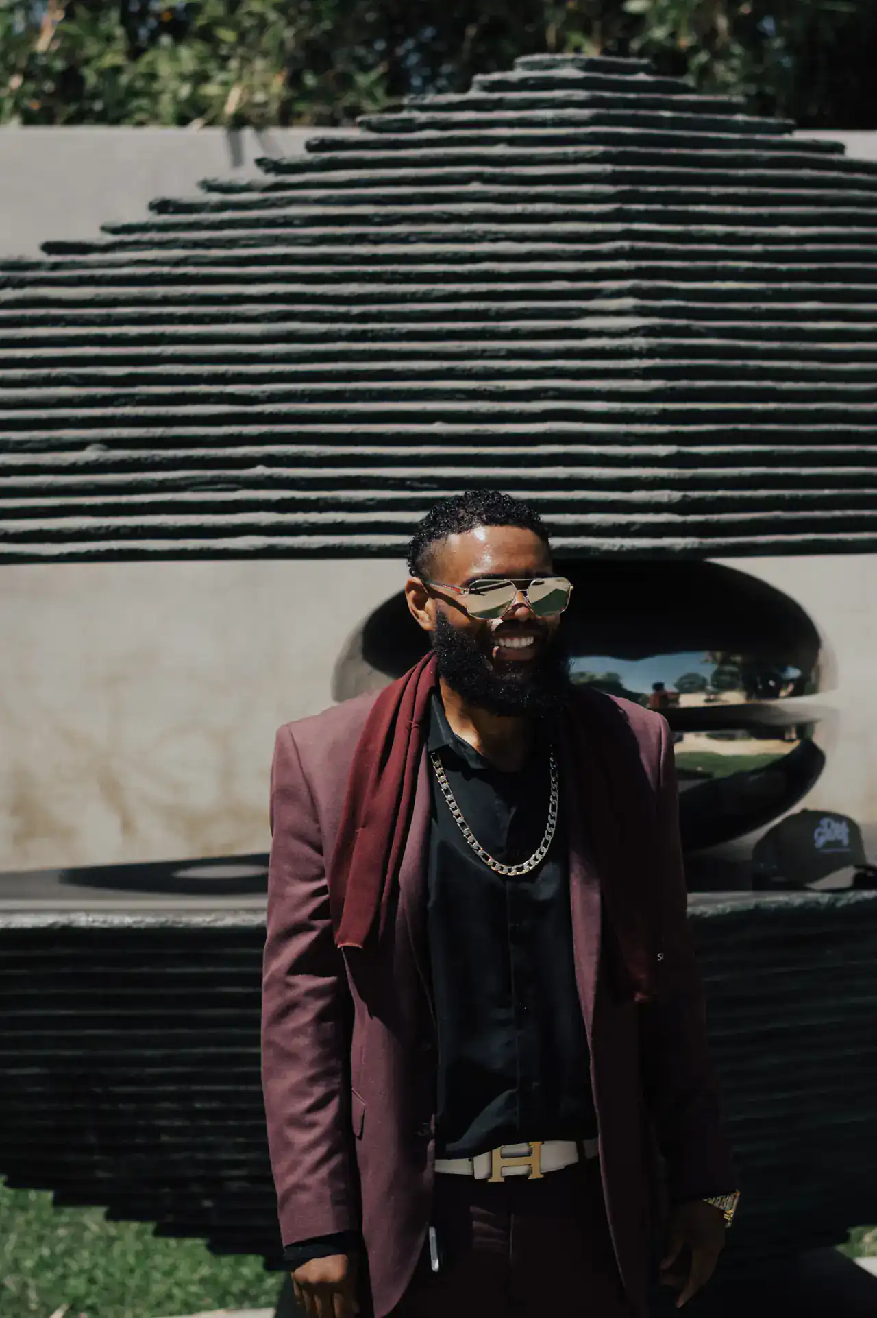 KingJu1c3 smiling in a burgundy blazer and gold chain in front of a stone sculpture